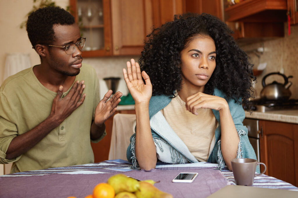 sad african american male in glasses holding hand on chest, apologizing to his angry and mad wife, who ignoring him, refusing to forgive him for cheating on her. people and relationships concept